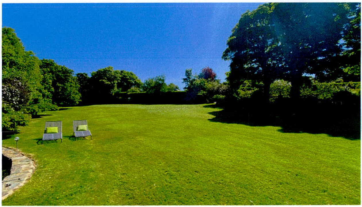 A sunny photograph showing a large, manicured green lawn surrounded by mature trees and shrubbery, with two sun loungers placed on the grass.