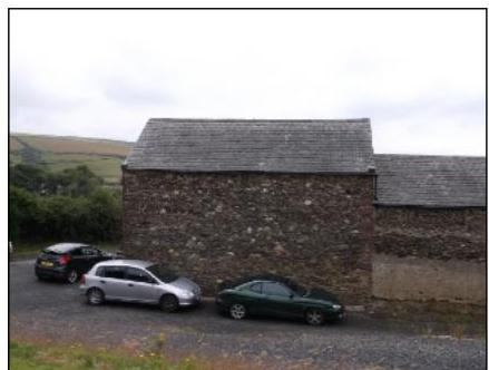 A photograph showing a stone building with a slate roof, likely a barn or outbuilding, with three cars parked on a gravel driveway in a rural setting.