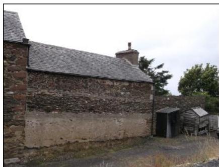 A photograph showing the side elevation of a stone building with a slate roof and chimney stack, alongside small wooden sheds.
