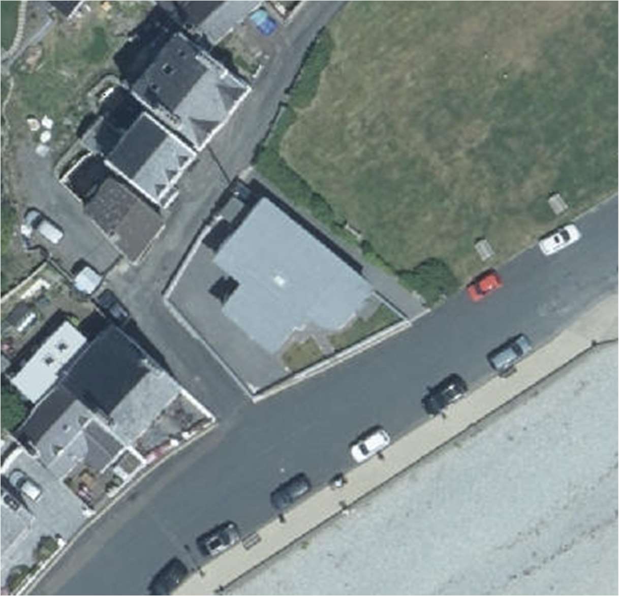 An aerial view showing a coastal site with a large flat-roofed building, adjacent houses, a road with parked cars, and a beach area.
