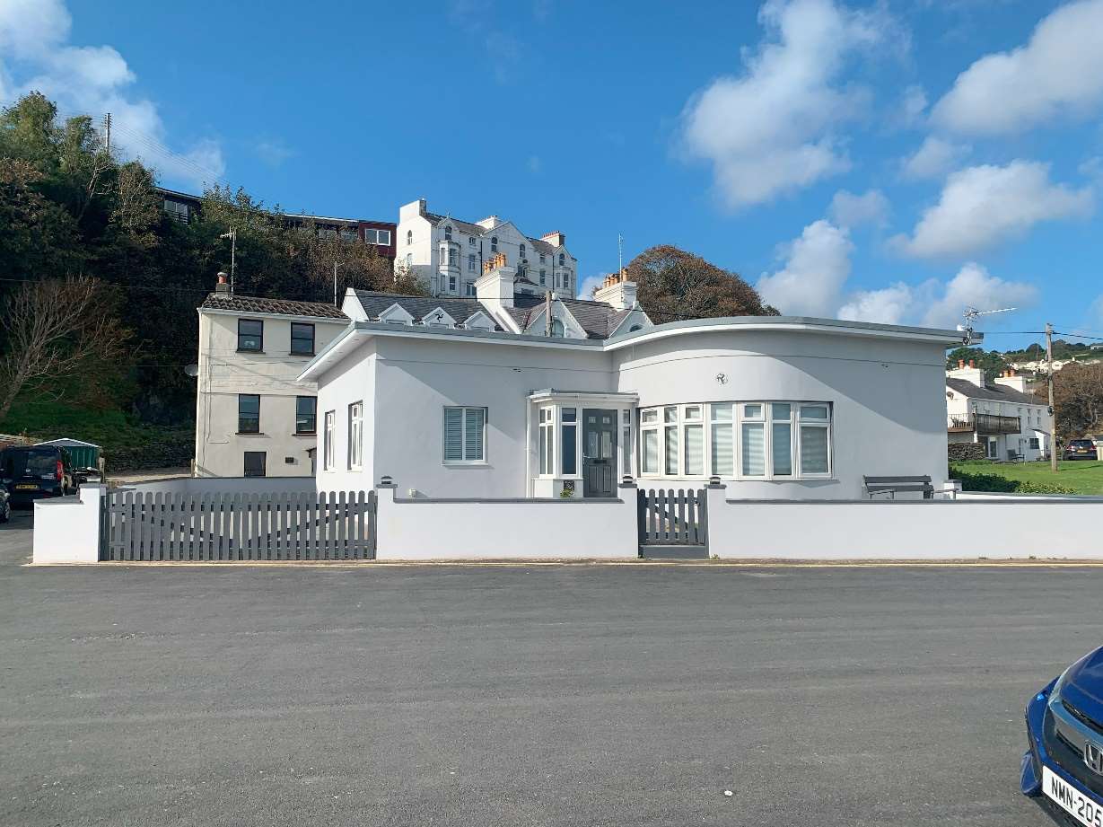 A street-level photograph of a white, single-story bungalow featuring a distinctive curved front section and dormer windows. The property is set back from the road behind a low white wall and grey picket fence, with a...
