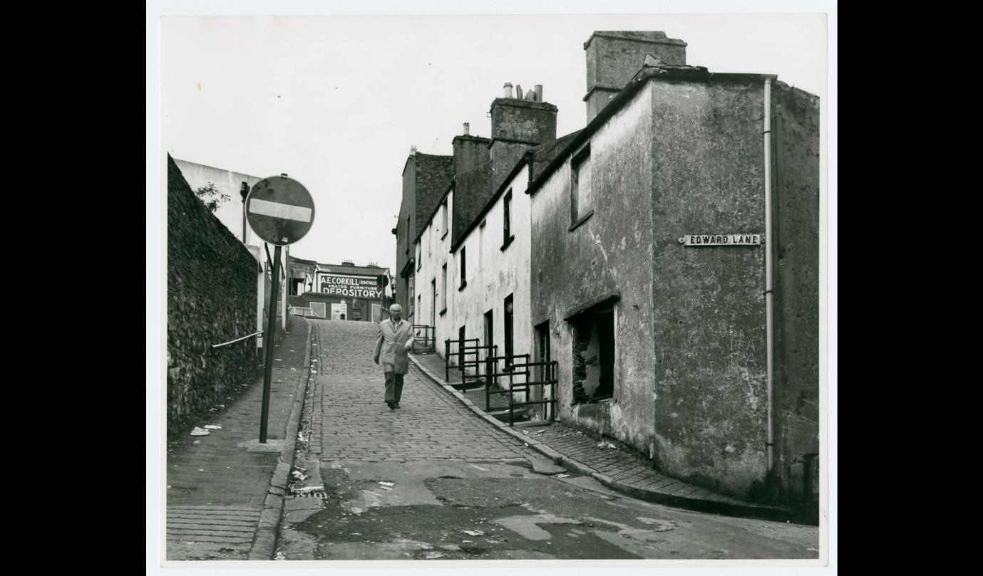 A black and white photograph showing a cobblestone street scene with a row of terraced buildings and a street sign reading 'Edward Lane'.
