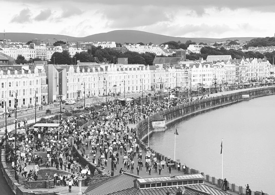 A black and white photograph showing a crowded promenade along a waterfront with large, uniform white buildings in the background.