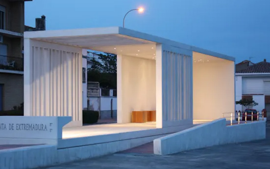 A photograph of a modern, illuminated white pavilion structure with vertical slats and a ramp at dusk.
