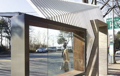 A photograph of a modern architectural structure with a white ribbed roof and glass walls, appearing to be a bus shelter or pavilion.