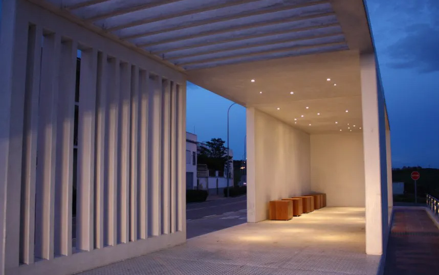 A photograph of a modern covered walkway or entrance structure featuring vertical slats, recessed lighting, and wooden benches at dusk.