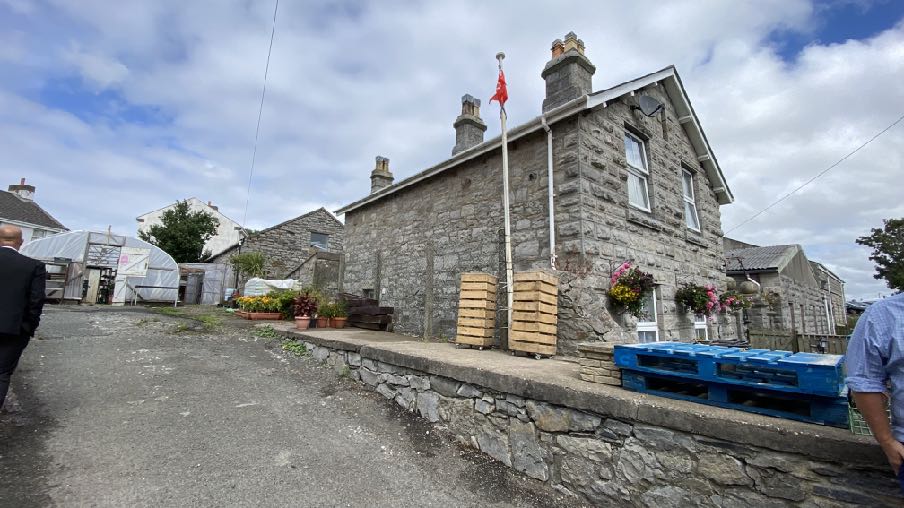 A photograph showing a stone building with a flag pole and stacked wooden pallets in the foreground, with a greenhouse visible in the background.