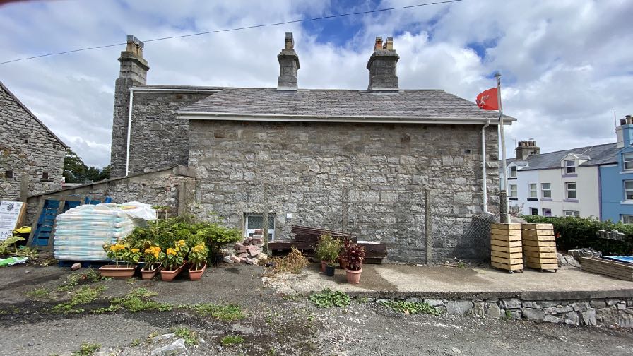 A photograph showing the exterior of a traditional stone building with slate roofing and chimneys, situated in a yard with potted plants and stacked materials.
