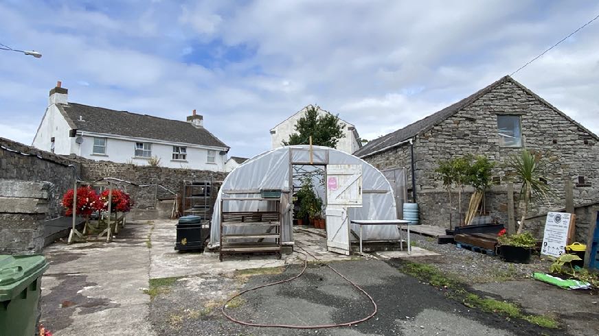 A photograph of an outdoor yard area featuring a white residential building on the left and a stone outbuilding on the right, with a polytunnel structure in the center.