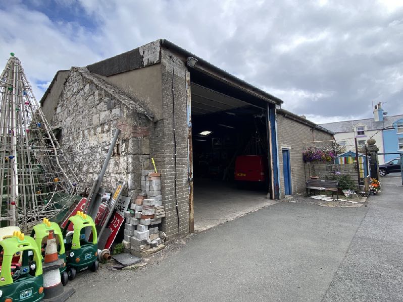 A photograph showing the exterior of an existing stone and brick workshop building with a large open bay door and paved yard.