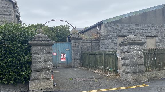 A street-level photograph showing the stone entrance pillars, a blue metal gate, and a large stone building with a wooden fence.