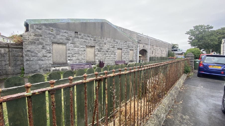 A photograph of a long, grey stone building with boarded-up windows, situated behind a rusty metal fence and wooden picket fence.