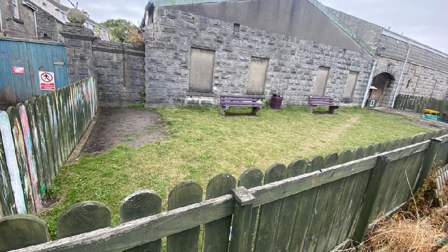 A photograph showing a grassy yard enclosed by wooden fencing, with a large stone industrial building featuring boarded-up windows in the background.