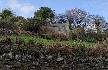 A photograph showing a stone house situated on a grassy slope, partially obscured by trees and bushes, with a dry stone wall in the foreground.