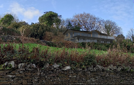 A photograph showing a two-story house situated on a grassy slope behind a dry stone wall, surrounded by trees and vegetation.