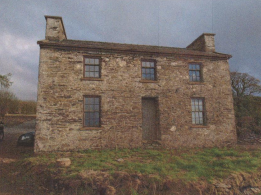 A photograph showing the existing two-story stone dwelling proposed for demolition, featuring a pitched roof and chimneys.