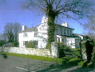 A photograph showing the existing white two-story dwelling with a stone boundary wall and driveway, set in a rural environment.
