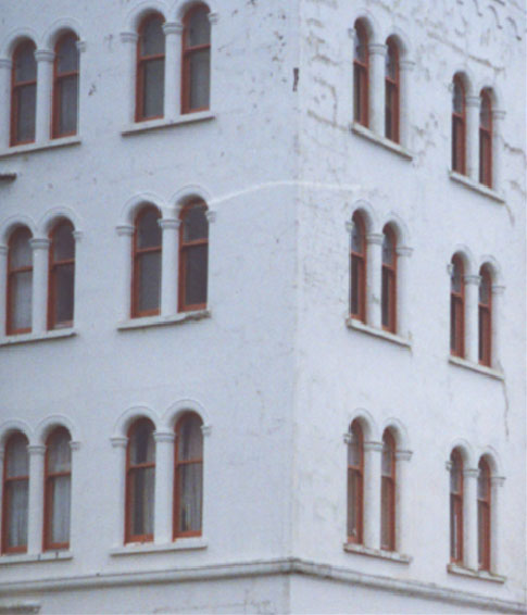 A close-up photograph of the corner of a white building featuring multiple rows of arched windows with reddish-brown frames.