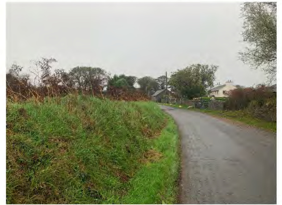 A street-level photograph showing a rural road curving through a grassy landscape with stone walls and houses in the distance.