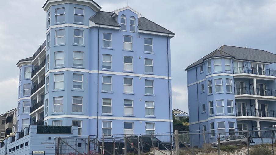 A photograph showing a large, multi-story light blue apartment building with balconies, situated next to a similar structure with a fence and parked cars in the foreground.