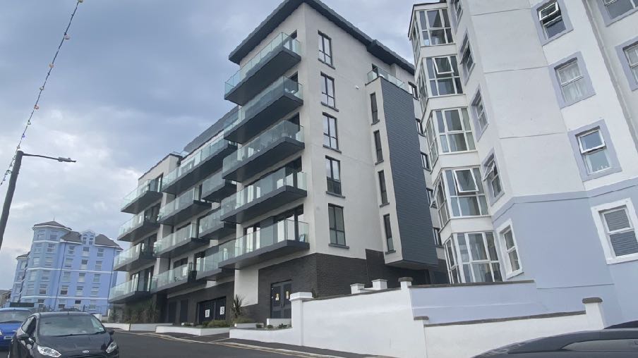 A low-angle photograph of a modern multi-story apartment block featuring glass balconies and white cladding. The building is situated on a street with a car and a white boundary wall visible in the foreground.