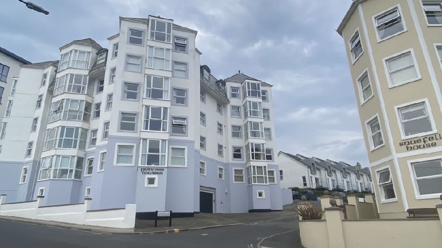 A street-level photograph showing a large white and blue apartment block named 'Princess Colleen' alongside other residential buildings in a coastal town setting.