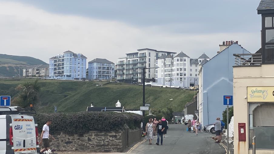 A street-level photograph showing a coastal town scene with large apartment blocks situated on a grassy hill in the background and pedestrians on the road below.