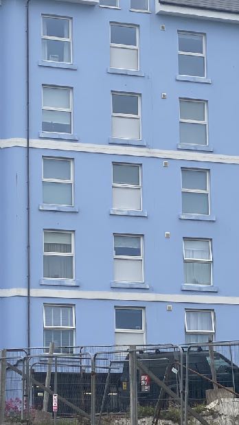 A photograph showing the blue exterior facade of a multi-story building with white window frames and a temporary construction fence in the foreground.
