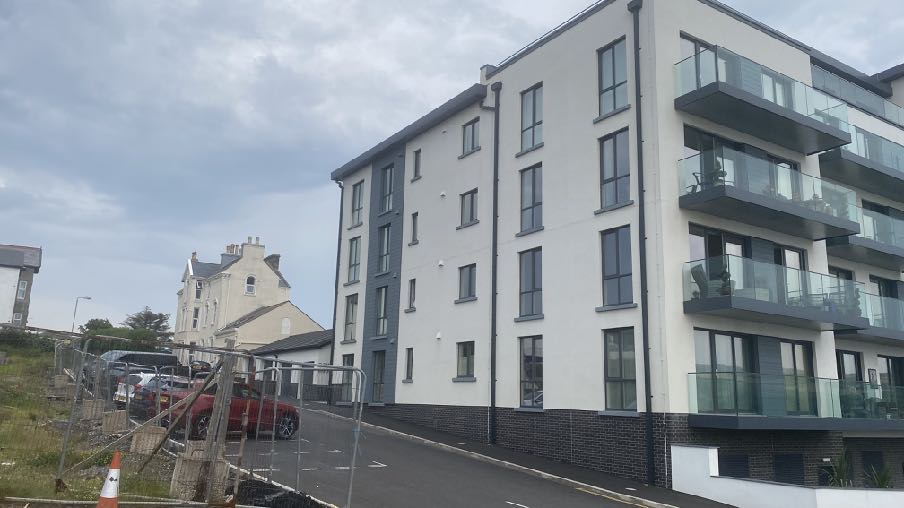 A photograph showing a modern multi-story apartment building with white and grey cladding and glass balconies, situated next to an older white house with construction fencing in the foreground.