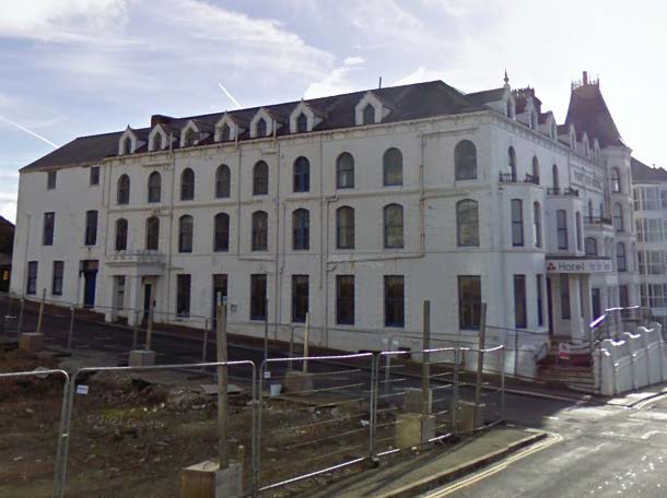 A photograph showing a large, white, multi-story building (resembling a hotel) with construction fencing and groundworks in the foreground.