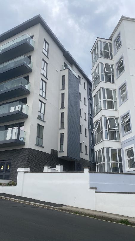 A street-level photograph showing modern multi-story apartment buildings with balconies and bay windows. A white boundary wall runs along the foreground in front of the development.