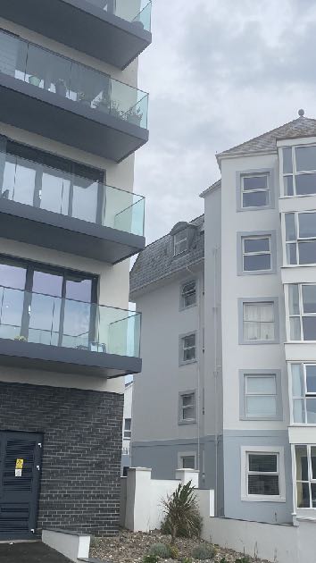 A street-level photograph showing a modern multi-story building with glass balconies and dark cladding adjacent to a traditional white building with a pitched roof. The foreground features a paved area with a small pl...