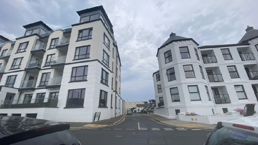 A street-level photograph showing two large, white multi-story apartment blocks flanking a road under a cloudy sky.