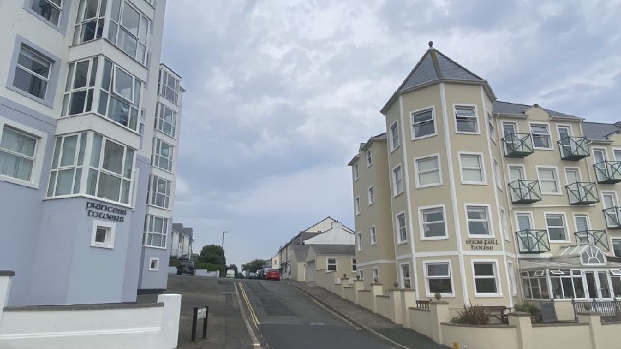A street-level photograph showing existing buildings on a sloping road, including a blue building labeled 'Princess Colours' on the left and a cream building with balconies labeled 'Spice Egg Delicacy' on the right.