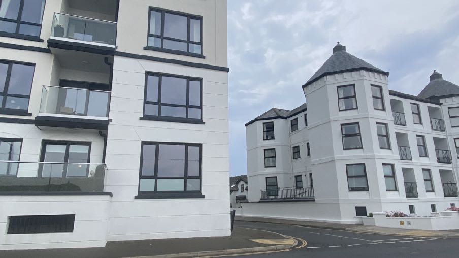A street-level photograph showing white residential buildings, including a modern block with balconies on the left and a building with a turret on the right.