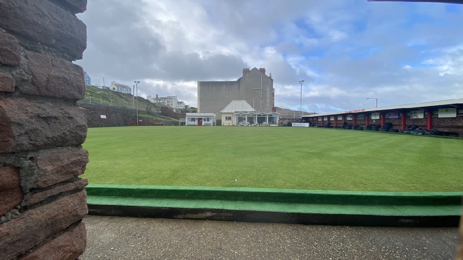 A photograph showing a large bowling green with a long clubhouse building on the right and a stone wall in the foreground.