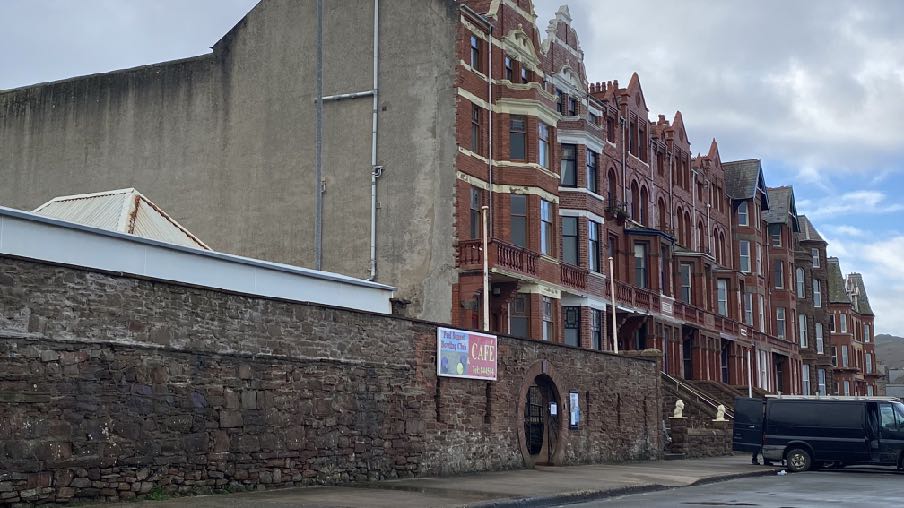 A street-level photograph showing a row of red brick terraced buildings next to a large concrete structure, separated by a stone wall with a cafe sign.