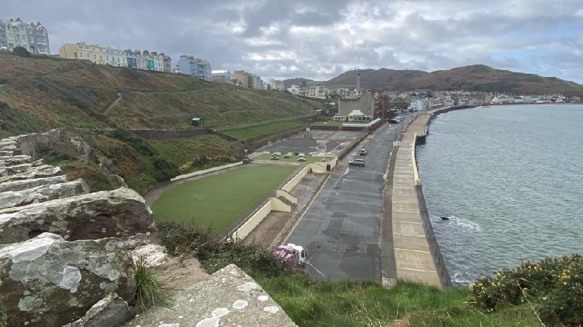 A high-angle photograph showing a coastal site in Peel, featuring old stone ruins in the foreground, a bowling green, a parking area, and a sea wall along the promenade.