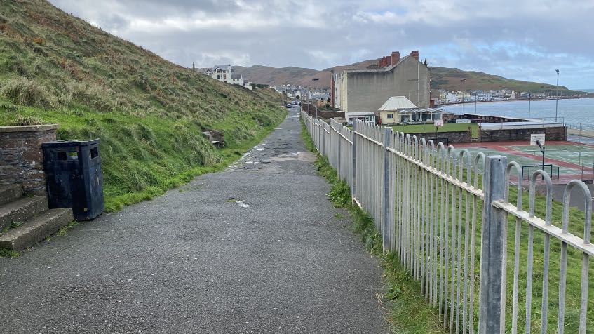 A photograph showing a paved pathway running alongside a grassy bank and metal fence, leading towards a coastal bowling facility and the sea.