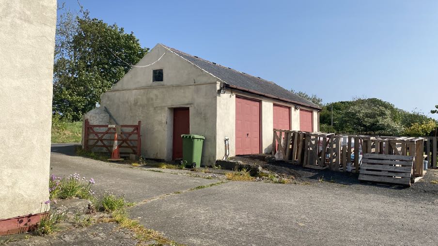 A photograph showing a detached, single-story building with a pitched roof and large red garage doors. The structure is situated on a paved area with wooden pallets stacked to the side under a clear blue sky.