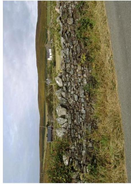 A rotated photograph showing a dry stone wall running alongside a road in a rural setting, with a white building visible in the distance on a hillside.