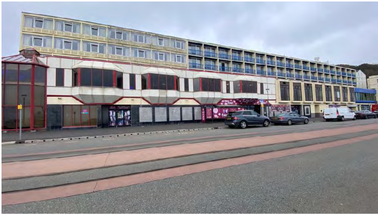 A street-level photograph showing the exterior of a large, multi-story commercial building, likely a hotel, with ground floor units and cars parked along the curb.