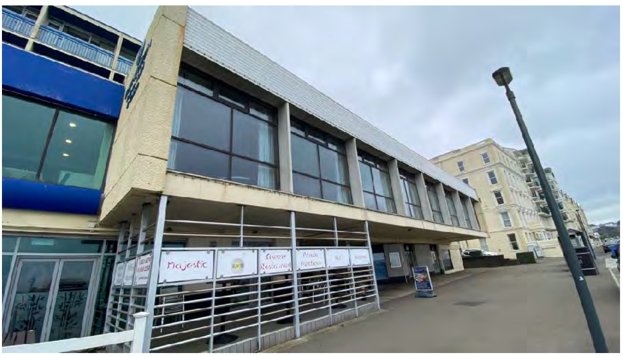 A street-level photograph of a commercial building, likely a hotel or bar, featuring a large ground-floor terrace enclosed by metal railings and a concrete upper facade.