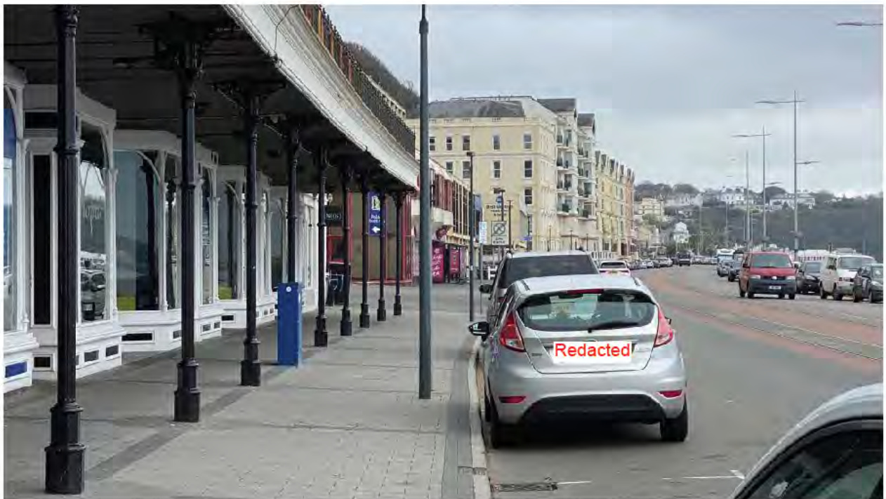 A street-level photograph showing a covered promenade with white shop fronts and black pillars on the left, alongside a road with parked and moving vehicles.