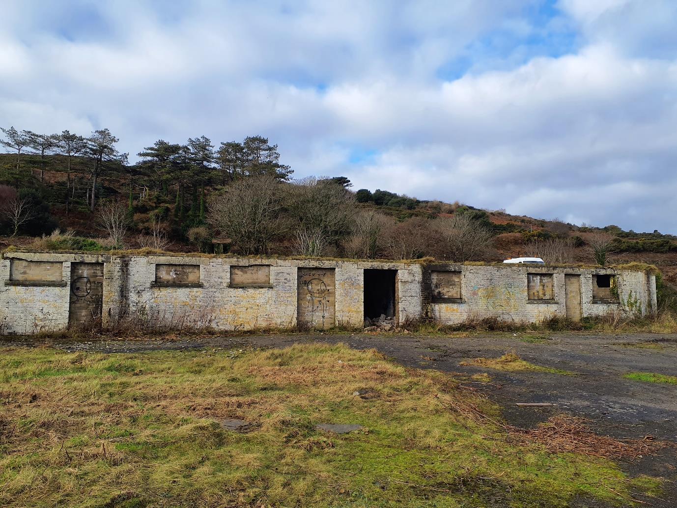 A photograph showing a long, derelict concrete building with multiple bays, possibly garages, situated in a grassy field with trees in the background.