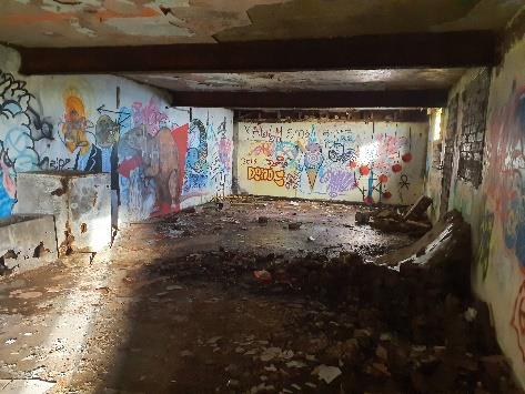 Interior photograph of a derelict concrete structure with extensive graffiti on the walls and debris scattered on the floor.