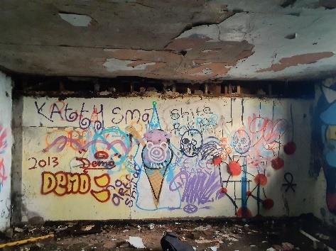 The image displays the interior of a derelict structure featuring a wall covered in colorful graffiti and a damaged, peeling ceiling.