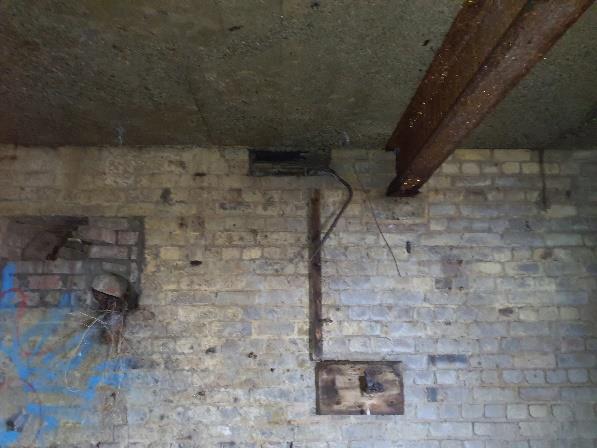 Interior photograph of a dilapidated brick wall with a concrete ceiling and rusty metal beam, likely part of a derelict structure.