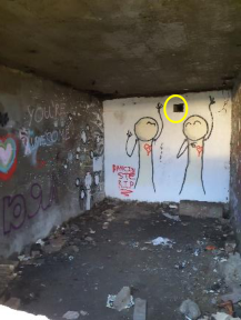 Interior photograph of a derelict room with graffiti-covered walls and debris on the floor, highlighting a small vent.
