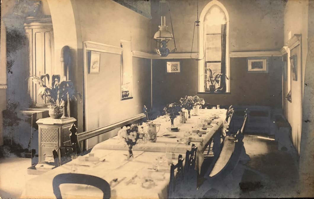 A vintage sepia-toned photograph showing a dining room interior with a long table, chairs, and a tall arched window.